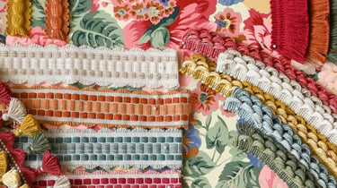A colourful arrangement of Sanderson passementerie trims displayed on a floral fabric background, featuring tassel fringes, braided cords, and woven borders in soft pastels and warm tones including coral, sage, mustard, and cream.