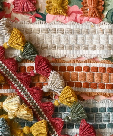 A colourful arrangement of Sanderson passementerie trims displayed on a floral fabric background, featuring tassel fringes, braided cords, and woven borders in soft pastels and warm tones including coral, sage, mustard, and cream.